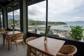 A restaurant with wooden tables and chairs, large windows offering a view of a body of water and mountains. - Tribhuwana Padma Lake Resort (Kintamani)