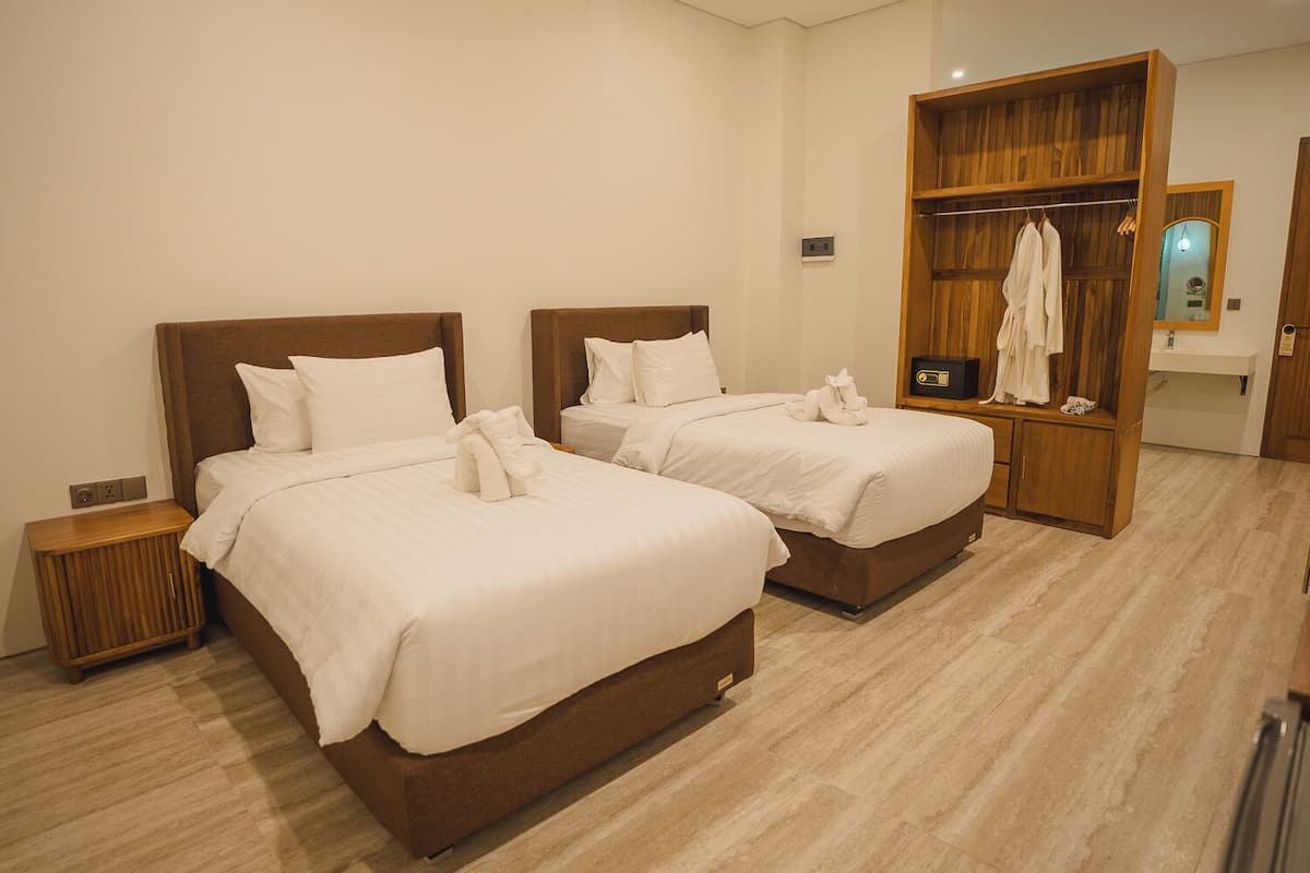 A hotel room with two beds, a wooden wardrobe, a bedside table, and a bathroom with a mirror and towel rack.