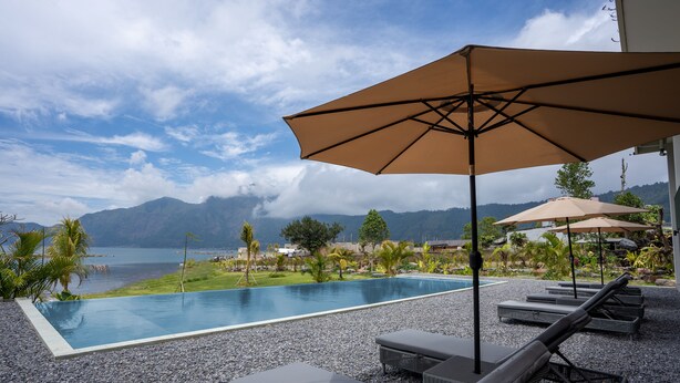 A poolside area with lounge chairs and umbrellas, overlooking a body of water and mountains. - Tribhuwana Padma Lake Resort (Kintamani)