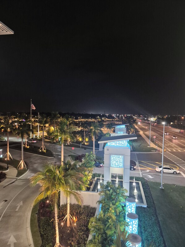 A nighttime view of a well-lit urban area with palm trees, a fountain, and a building with illuminated blue panels.