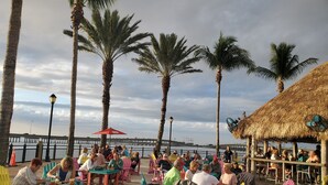 A beachside dining area with tables and chairs, palm trees, and a thatched-roof structure. - TROPICAL WATERFRONT/POOL  BEACH, BOAT🛥 HOME BY VENICE SIESTA KEYS BEACH PETS OK (Port Charlotte)