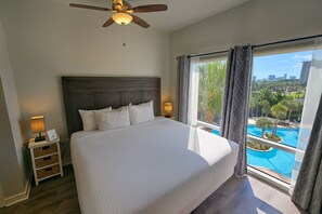 A bedroom with a large bed, a ceiling fan, a nightstand with a lamp, and a window with a view of a swimming pool and buildings. - Luau 6424 | 1BR Condo w/ Gulf Views, Pool Views & Steps to the Beach (Miramar Beach)