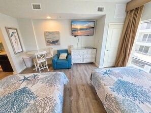 Une chambre d’hôtel avec deux lits, un bureau, une chaise, une télévision et un tableau représentant une scène de plage.