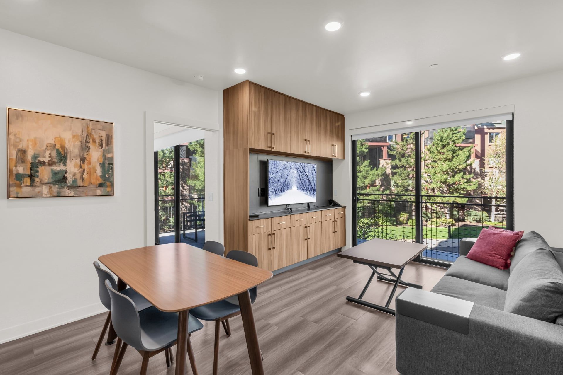 A modern living space with a dining area, a sofa, a TV, and a view of a green outdoor area.