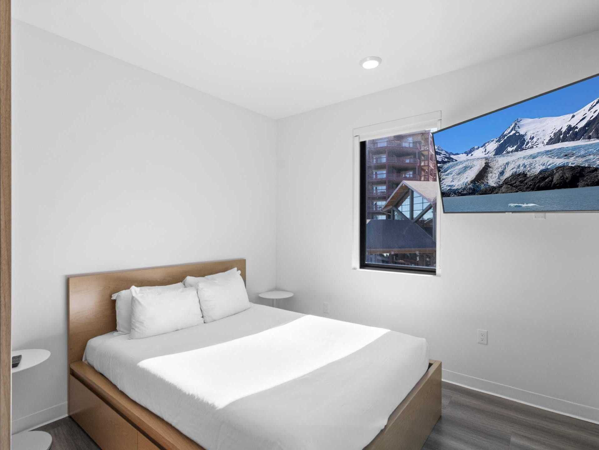A modern bedroom with a bed, a small bedside table, a window with a view of mountains and a building, and a wall-mounted TV.