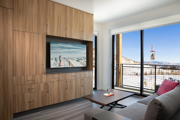 Een moderne woonkamer met een grote flatscreen televisie bevestigd aan houten kasten, een bank met een rood kussen en uitzicht op een bergachtig landschap door grote ramen.