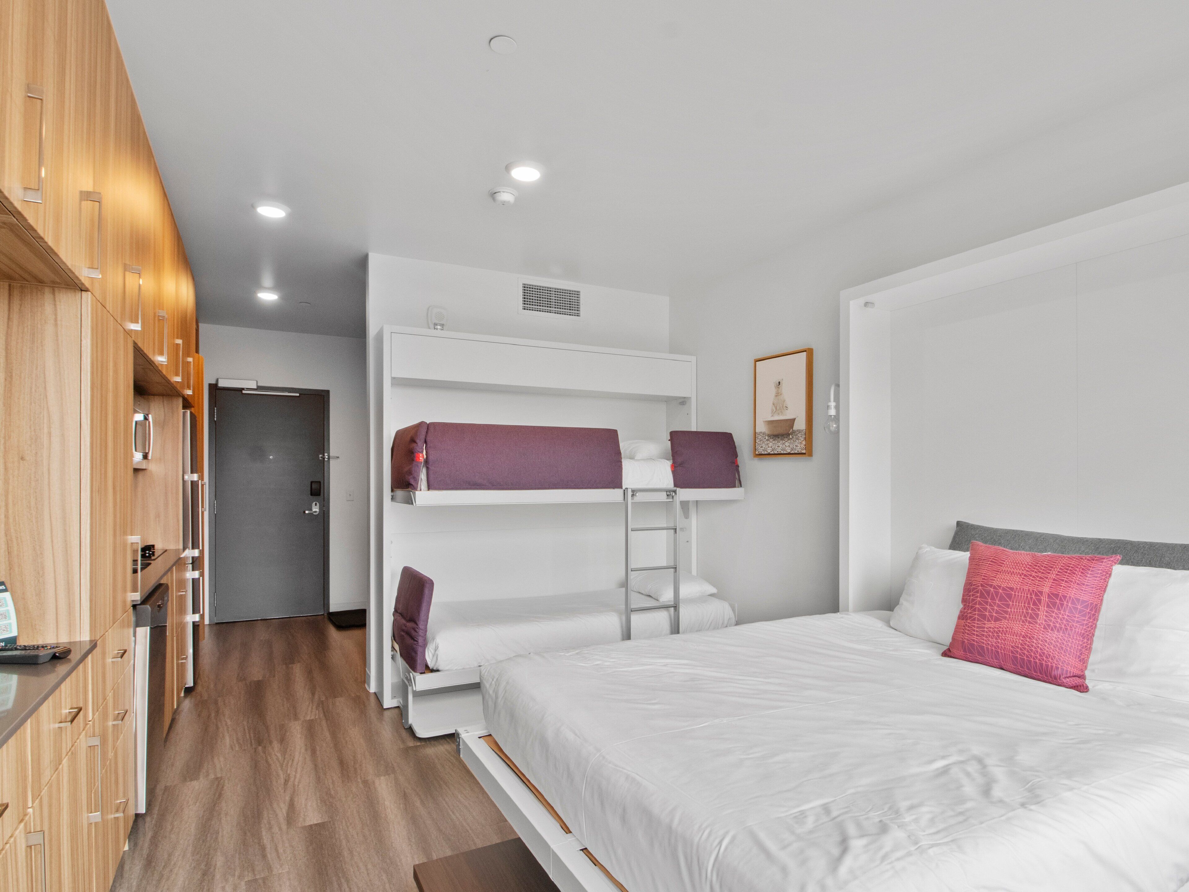 Une chambre d’hôtel moderne équipée d’un lit superposé, d’un lit séparé, de meubles en bois et d’une kitchenette.