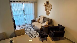 A modern living room with a sofa, a dining table, and a coffee table. There is a large window with curtains, a wall-mounted air conditioner, and a decorative wall piece. - Luxury Celebration Apartment - Belém, PA (Belém)