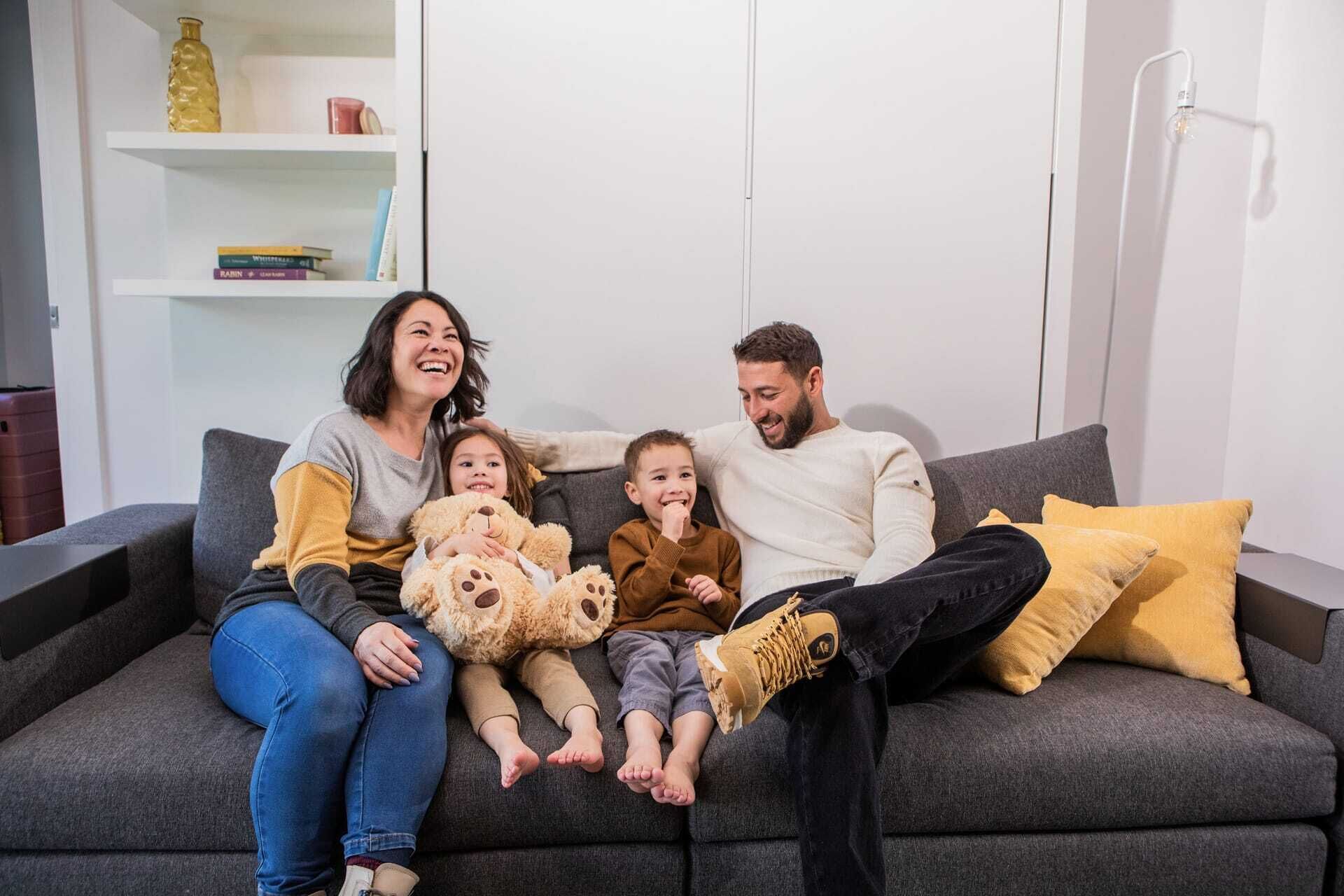 En familie sidder på en grå sofa med en bogreol og en lampe i baggrunden.