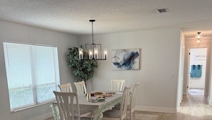 A dining area with a wooden table, white chairs, a chandelier, and a large window with blinds.