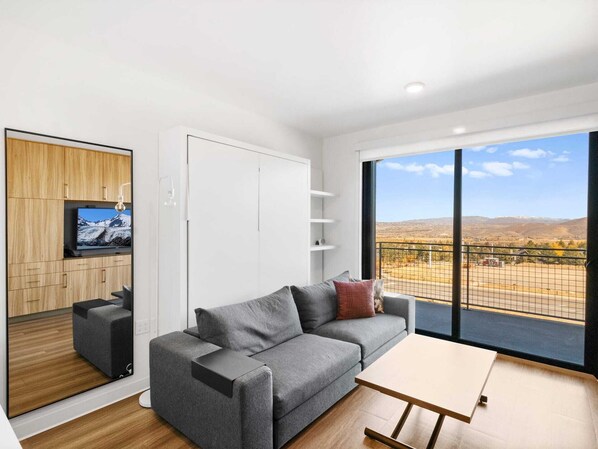 A modern living room with a grey sofa, a small table, a TV mounted on a wooden cabinet, and a large window with a view of mountains.