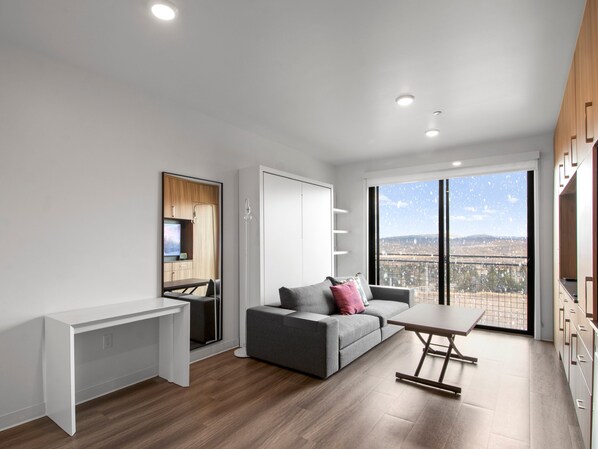 Um apartamento moderno com sofá cinzento, secretária branca e mesa de jantar. Apresenta uma janela ampla com vista para a cidade.