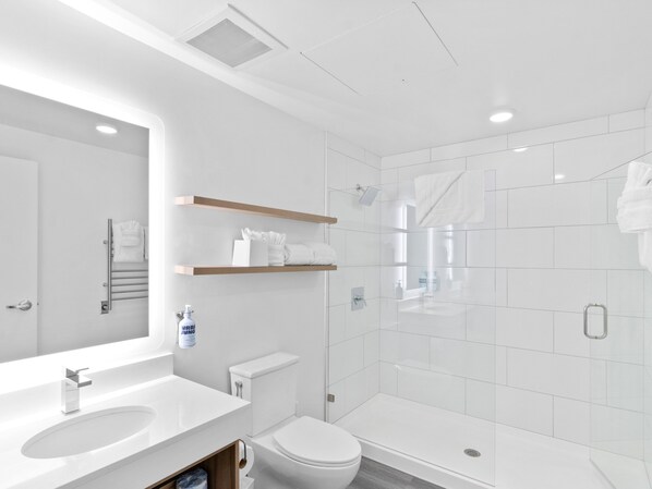 A modern bathroom with a white sink, toilet, and shower area. There are wooden shelves above the toilet and a large mirror above the sink.