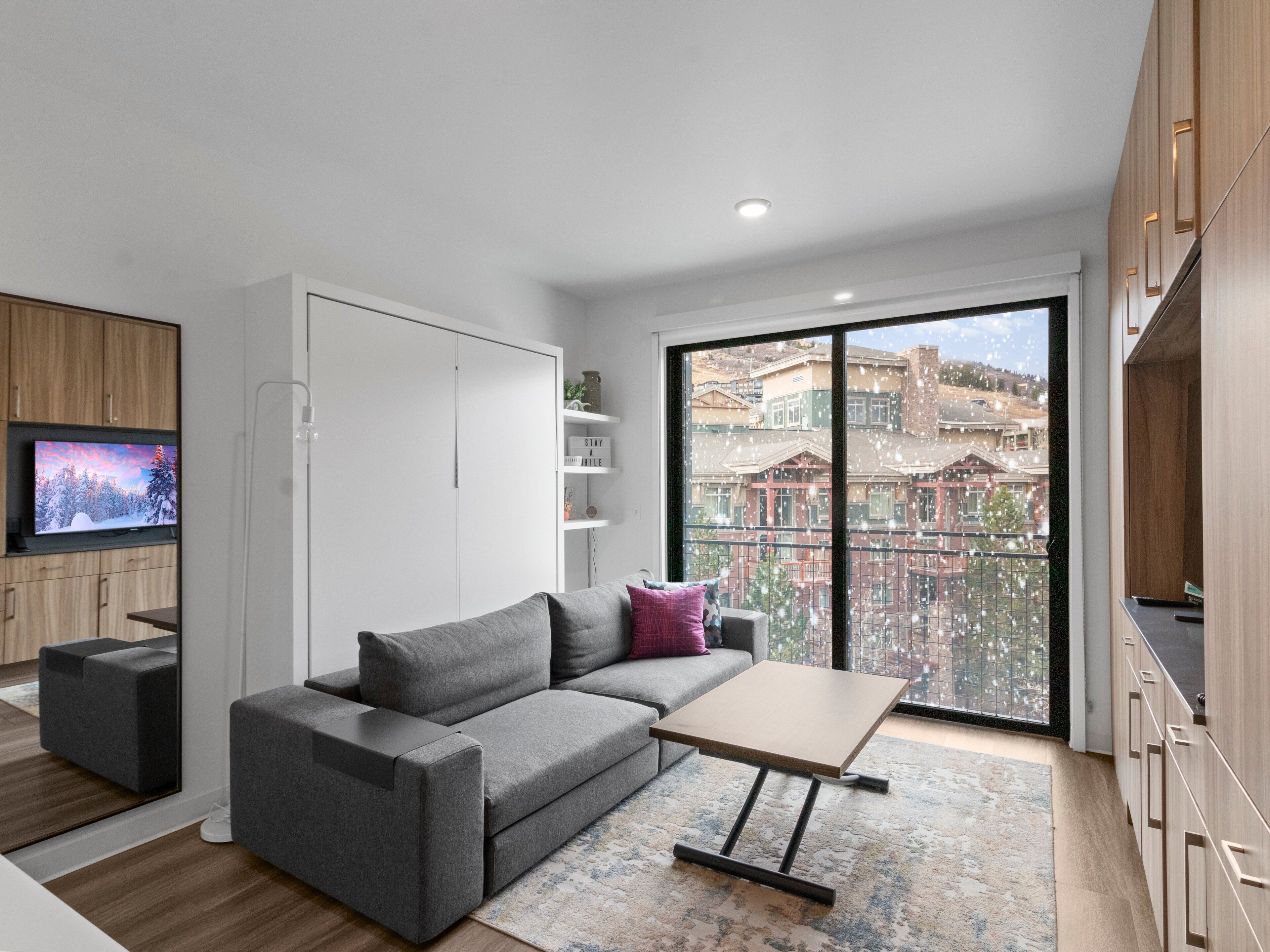 Un salon moderne avec un canapé gris, une petite table et un téléviseur posé sur un meuble en bois. Une grande fenêtre offre une vue sur les immebles et donne accès à un balcon.