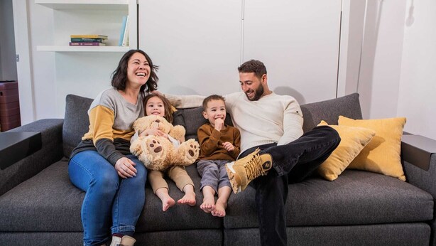 Eine Familie sitzt auf einem grauen Sofa, im Hintergrund sind ein Bücherregal und eine Lampe zu sehen.