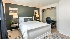 Habitación de hotel con cama, escritorio y silla. Hay una ventana con cortinas, una lámpara y una pequeña planta en la mesita de noche.