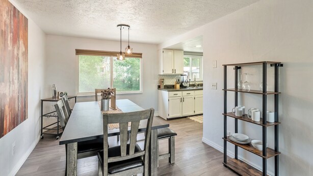 Un comedor con mesa y sillas de madera, una lámpara colgante y una cocina al fondo.