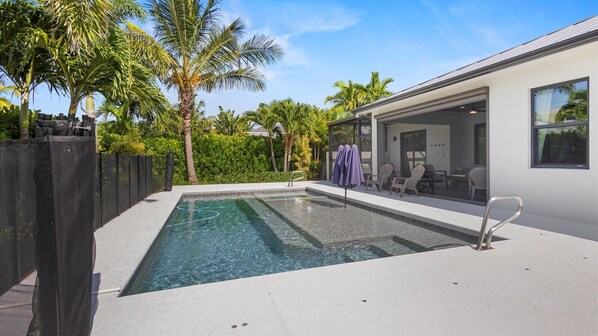 A modern outdoor pool area with a clear pool, surrounded by palm trees and a covered seating area with chairs.