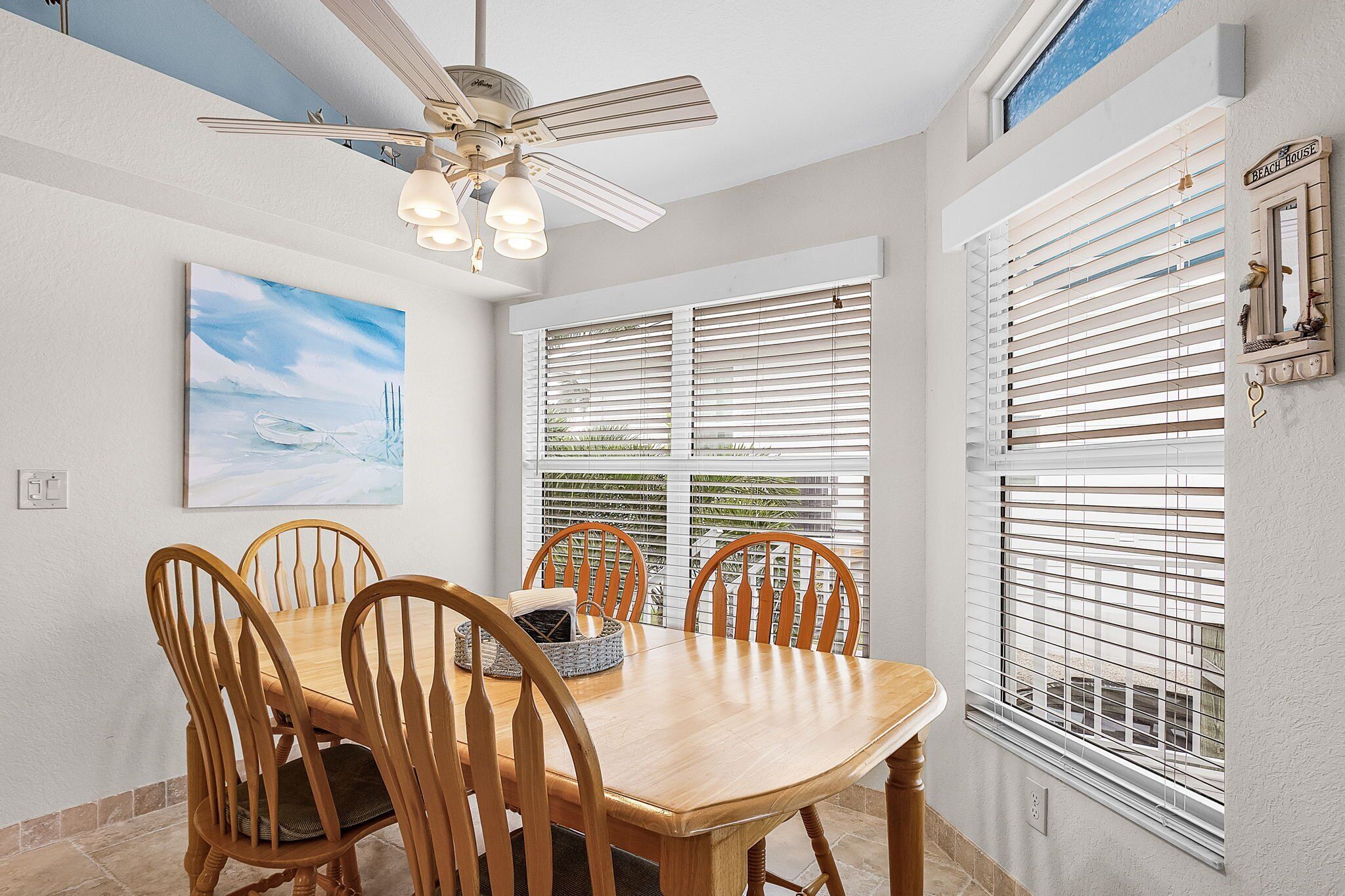 A dining area with a wooden table and chairs, a ceiling fan, and a painting on the wall.