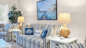 Een woonkamer met gestreepte banken, een groot ingelijst schilderij van een gracht, twee tafellampen en een potplant.