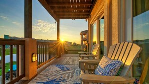 A covered balcony with a patterned rug, wooden chairs, and a view of the sunset. - 2 Homes, Sleeps 22 Steps to the Beach, Private Pool, Fire Pit, Balcony Views (Port Aransas)