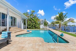 Ein rechteckiger Pool mit gewölbter Kante, umgeben von einer Ziegelterrasse. Liegestühle und eine Bank mit gestreiften Handtüchern sind vorhanden. Im Hintergrund sind Palmen und ein Gebäude mit großen Fenstern zu sehen.