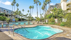 A swimming pool with a deck, surrounded by palm trees and a building with balconies.