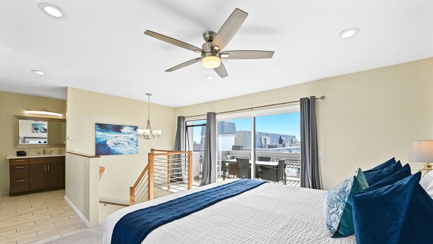 A hotel room with a bed, a ceiling fan, a large window, and a view of the city.
