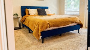 A bedroom with a bed, a nightstand, a window with blinds, and a painting on the wall.
