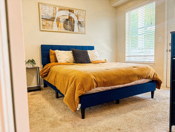 Un dormitorio con cama, mesita de noche, ventana con persianas y un cuadro en la pared.