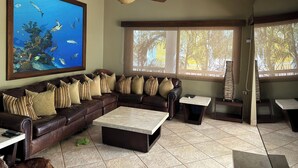 Una sala de estar con un sofá de cuero marrón, un mural grande de peces, un ventilador de techo y vistas a palmeras a través de las ventanas.