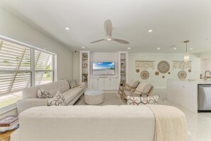 Ein modernes Wohnzimmer mit Sofa, Hocker und Sesseln, einem Deckenventilator und einem Fernseher an einer weißen Wand.