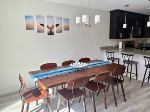 A dining area with a wooden table, six chairs, a blue runner, and a whale-themed wall art.
