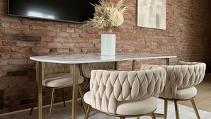 A dining area with a brick wall, a flat-screen TV mounted above a round table, and two upholstered chairs with a tufted design.