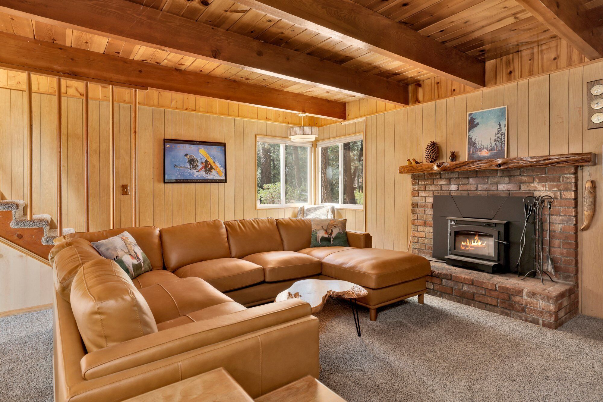 Una sala de estar acogedora con un sofá seccional de cuero, chimenea, paredes de madera y un ventanal con vistas a árboles.