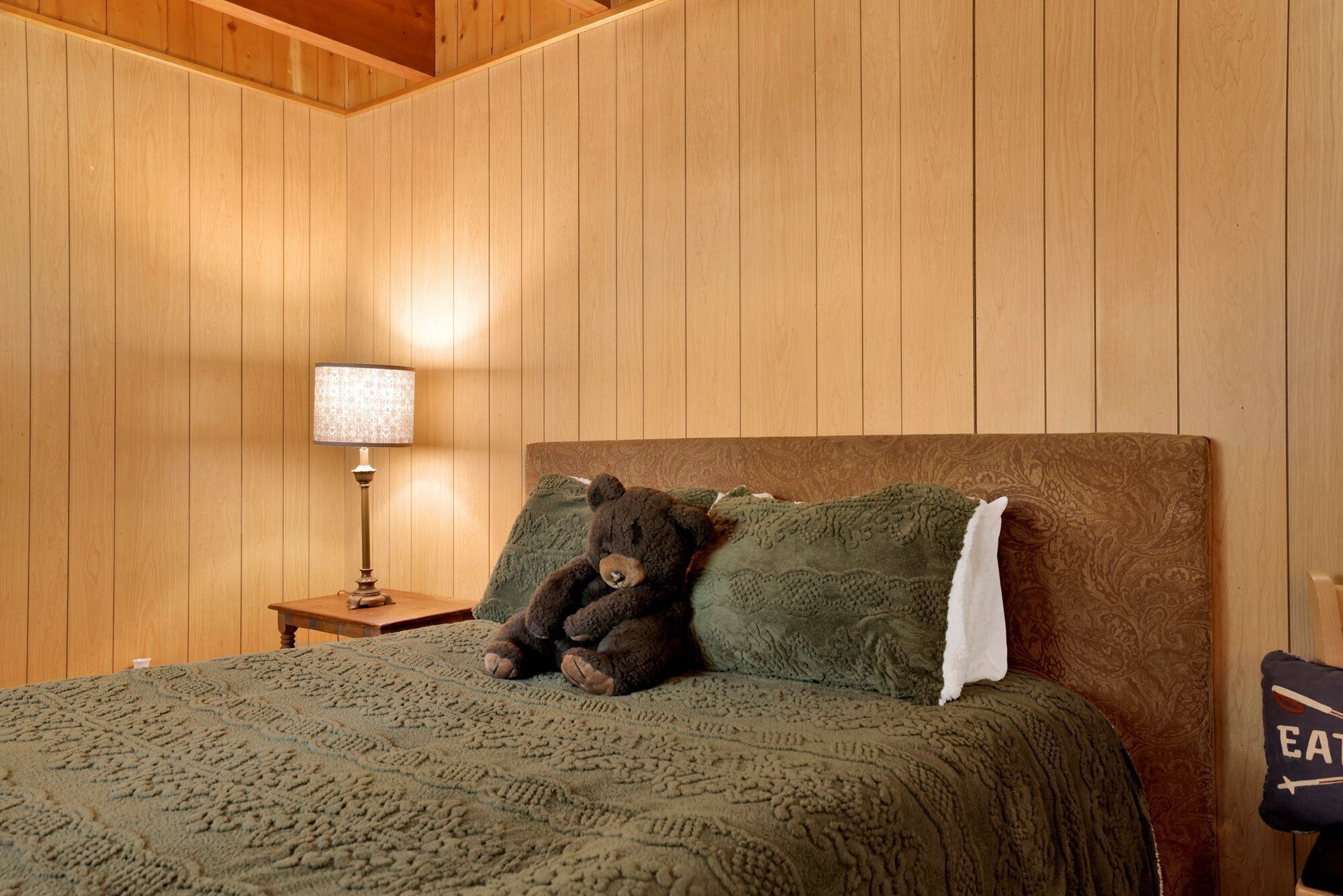 Une pièce aux murs lambrés, avec un lit, une lampe et un ours en peluche posé sur le lit.