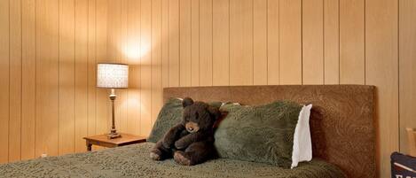 Une pièce aux murs lambrés, avec un lit, une lampe et un ours en peluche posé sur le lit.