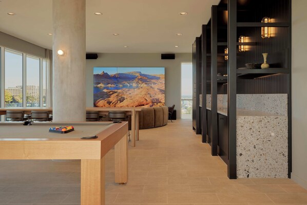 A modern hotel lobby with a pool table, seating area, and a large wall mural of a desert landscape.