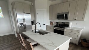A modern kitchen with a central island, wooden chairs, a stainless steel refrigerator, and a microwave. - Heart of Homer - Redoubt (Homer)