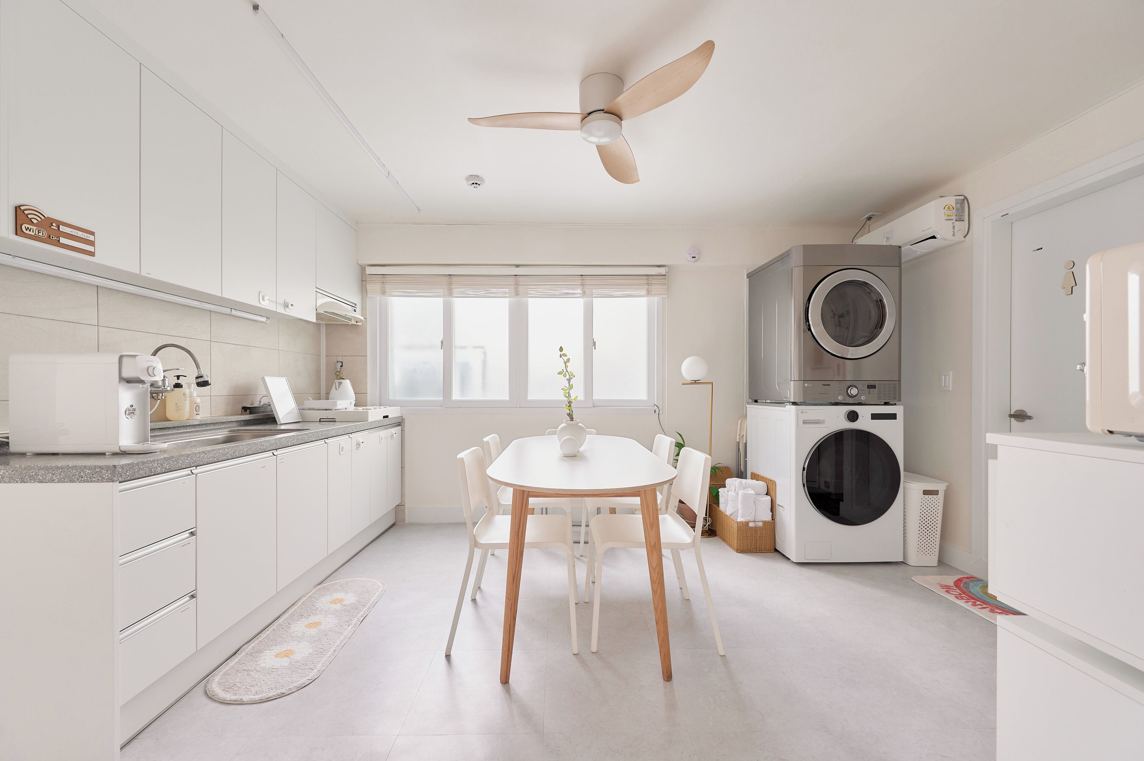A modern kitchen with white cabinets, a dining table, chairs, a ceiling fan, a washing machine, and a large window.