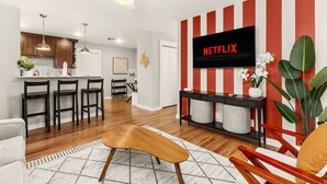 Una sala moderna con un televisor de pantalla plana montado en una pared a rayas rojas y blancas, una mesa de centro de madera y una cocina al fondo.