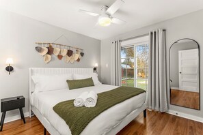 Una cama bien tendida con una manta verde y toallas enrolladas, un ventilador de techo, una lámpara de pared y un amplio ventanal con cortinas.