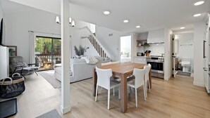A modern living room with a wooden dining table, white chairs, a grey sofa, and a staircase leading to an upper level.