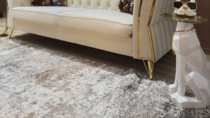 Ein weißes, gestepptes Sofa mit goldenen Beinen, ein dekorativer Tisch mit einer weißen Katzenfigur und ein gemustertes Teppich in einem Raum mit braunen Wänden und einem großen Fenster.