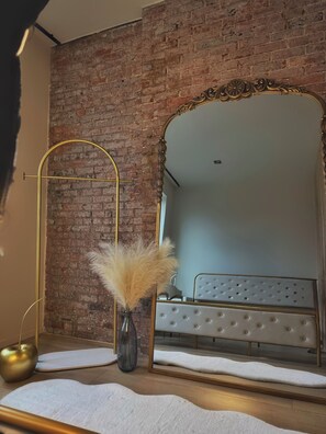 Ein Schlafzimmer mit Backsteinwand, einem großen Spiegel, einem Bett mit gestepptem Kopfteil und einem goldgerahmten Schminktisch.