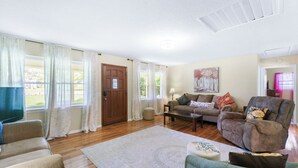 A spacious living room with a wooden floor, a large window with curtains, a sofa, a chair, and a painting on the wall. - Lookout Mtn. Escape w/ Fire Pit & 1 Acre Level Lawn- Mins to Cloudland Canyon (Rising Fawn)