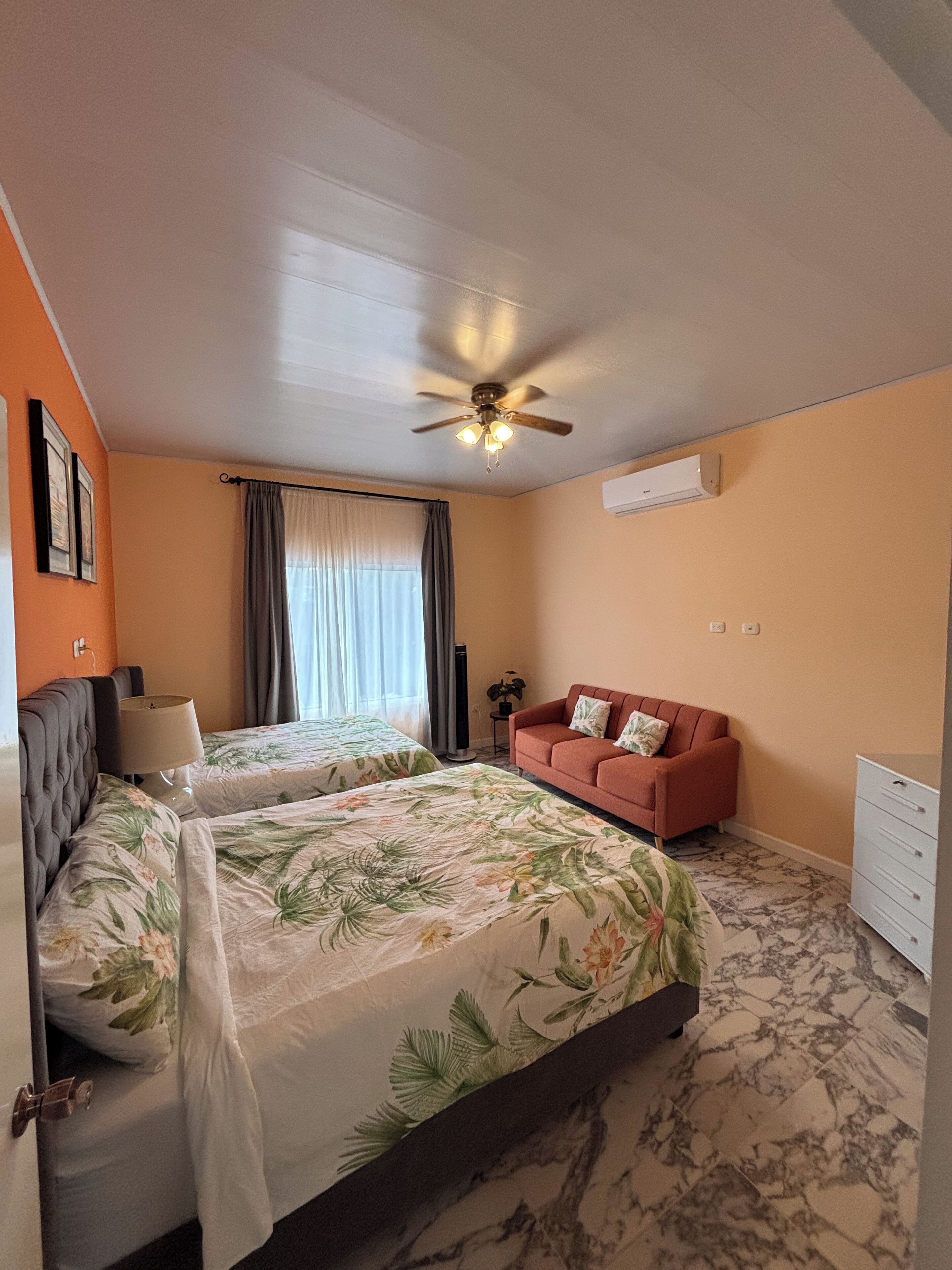 Een hotelkamer met twee bedden, een slaapbank, een plafondventilator en een marmeren vloer.