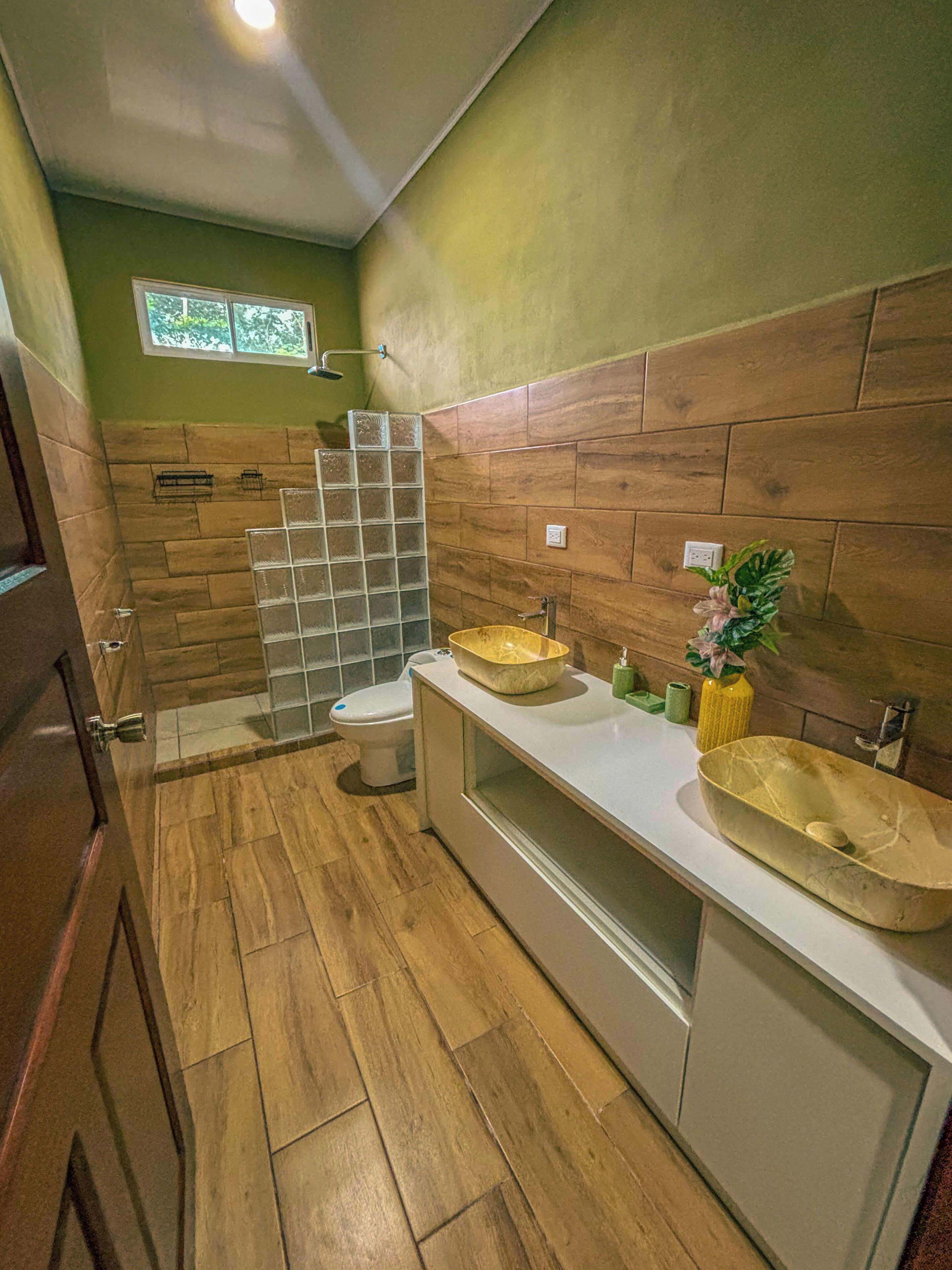 Een badkamer met houten vloer, toilet, douchecabine van glas en twee wastafels met een plantje op het aanrecht.