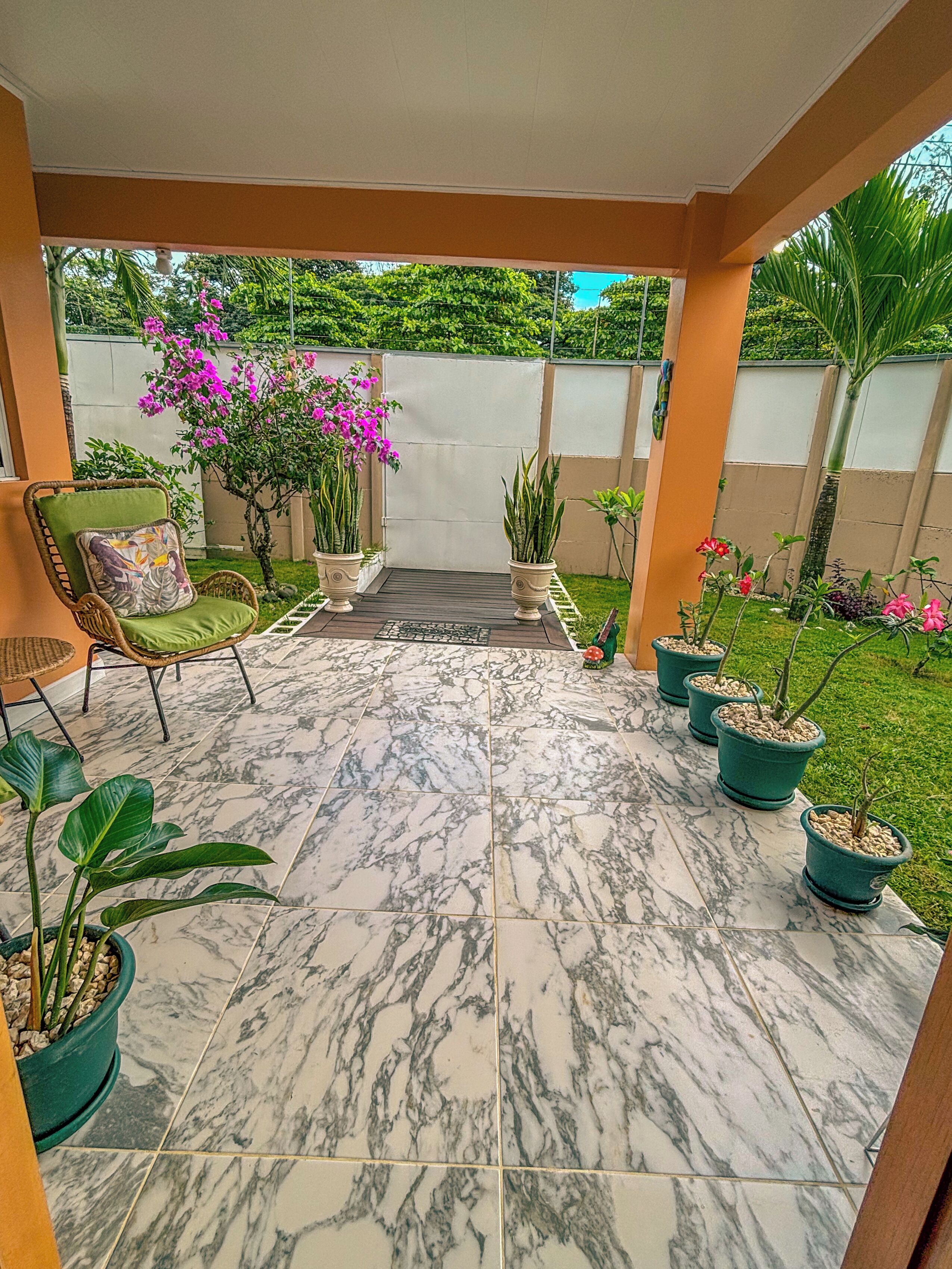 A covered patio area with a tiled floor, outdoor furniture, potted plants, and a view of a lush garden.