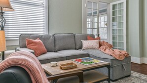 A living room with a gray sectional sofa, a coffee table, and a lamp. There is a window with blinds and a door with glass panels. - Cozy 2nd-Floor Suite near Beach, The Strand, Pier (Galveston)
